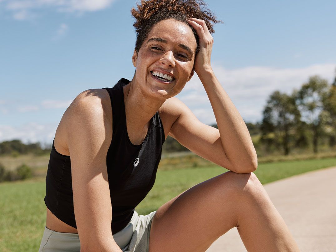 A woman smiles and looks off to the side, sitting outside on a sunny day wearing a black crop top and grey shorts